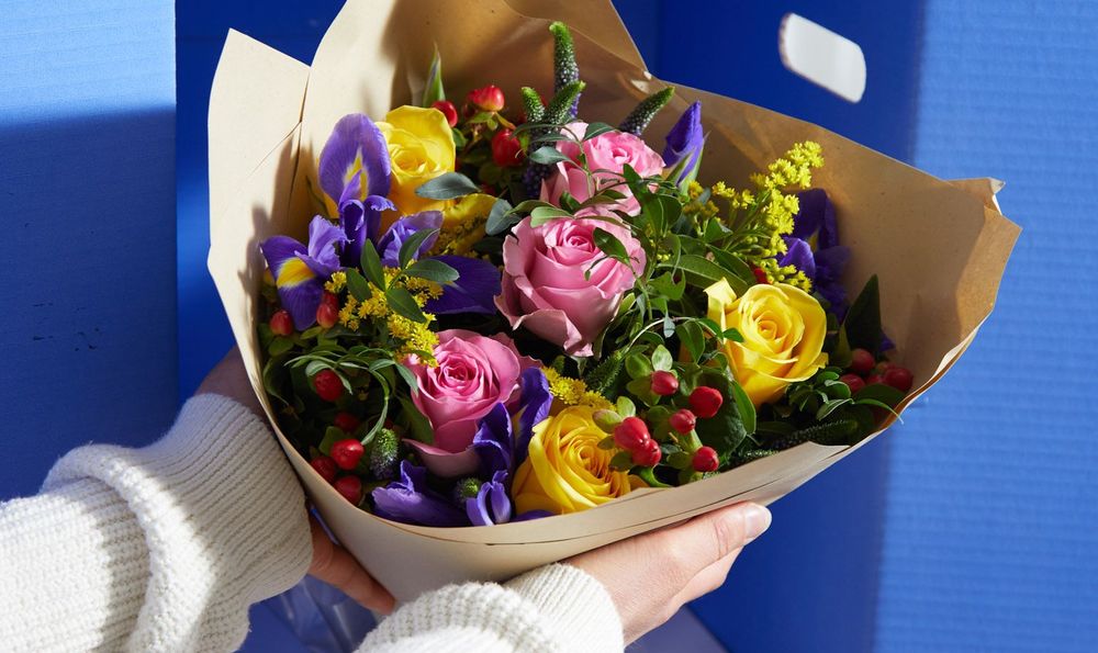 A bouquet of pink and yellow roses, red berries and purple purple veronica are being lifted out of a a box